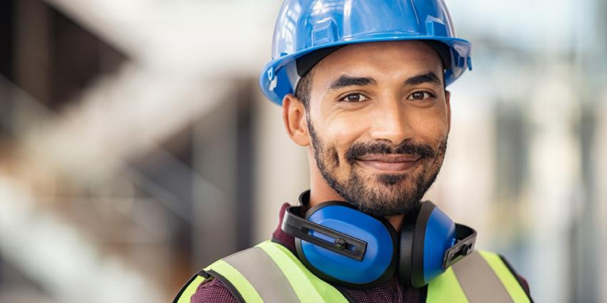 The image shows a construction worker wearing a blue hard hat, high-visibility vest, and protective earmuffs around his neck.