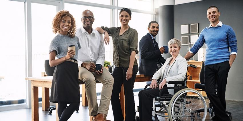 The image shows a diverse group of professionals standing and sitting together in a modern office setting. The group includes individuals of different ages, genders, and one person in a wheelchair, highlighting inclusivity and accessibility. They are all smiling and appear relaxed, suggesting a positive and collaborative work environment, likely involved in a consultation or team-building session.