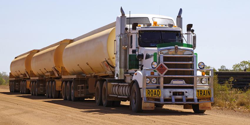 The image shows a large "road train" truck transporting multiple tanker trailers on a dirt road, marked with a hazardous materials sign