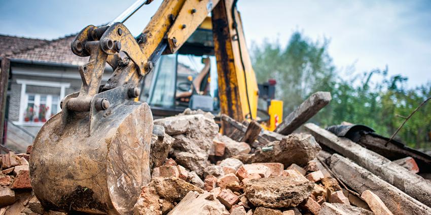 The image shows an excavator bucket amidst rubble, breaking down bricks and concrete, highlighting an active demolition site.