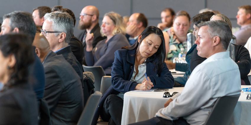 The image shows a group of people seated at tables, attending an event or conference, with one person taking notes