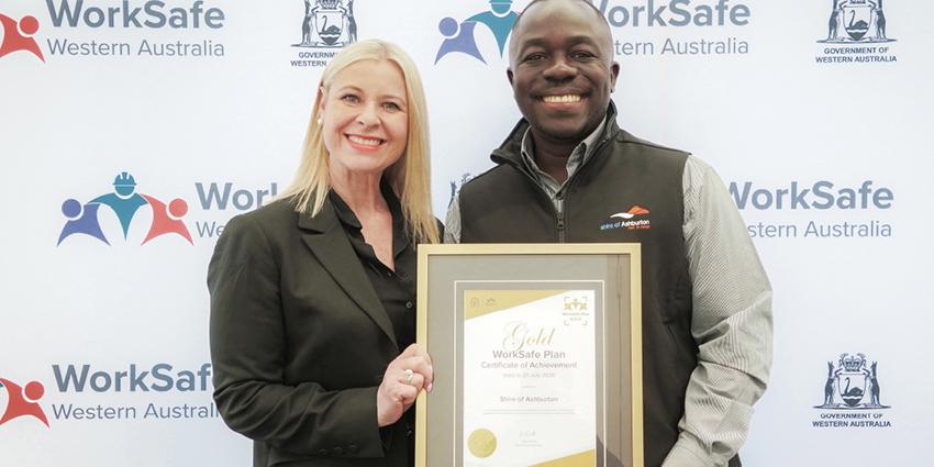The image shows two people standing in front of a WorkSafe Western Australia banner, holding a "Gold WorkSafe Plan Certificate of Achievement"